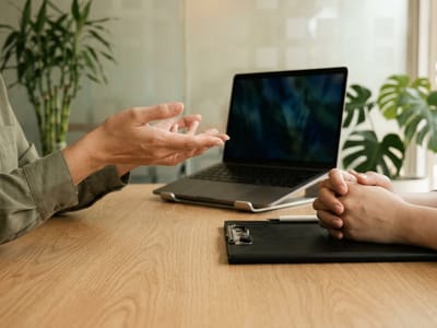 Two people seated at a table with a laptop, one gesturing and the other with folded hands