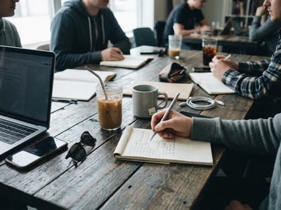 People around a wooden cafe table with notebooks, laptop and drinks while one writes in a notepad